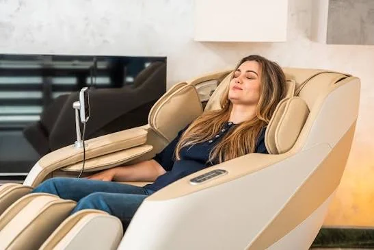 Beige and cream full-length massage chair shown fully reclined in a living room, with a person resting and a tablet-style controller on a stand beside the armrest